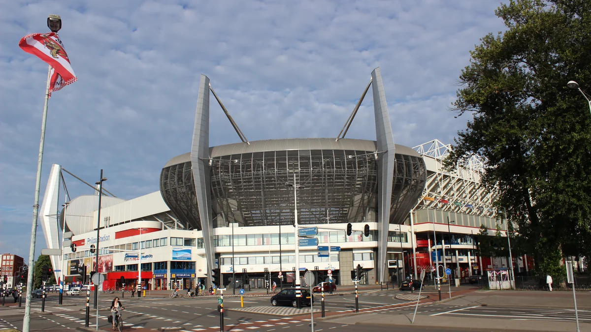 Philips Stadion visto de um cruzamento, com mastros, árvores, ciclistas e carros