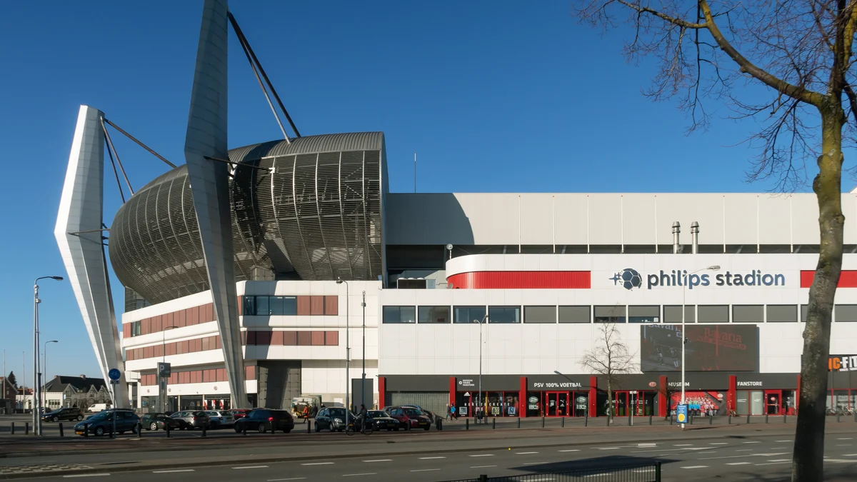 Fachada branca do Philips Stadion sob céu azul, com carros estacionados em frente