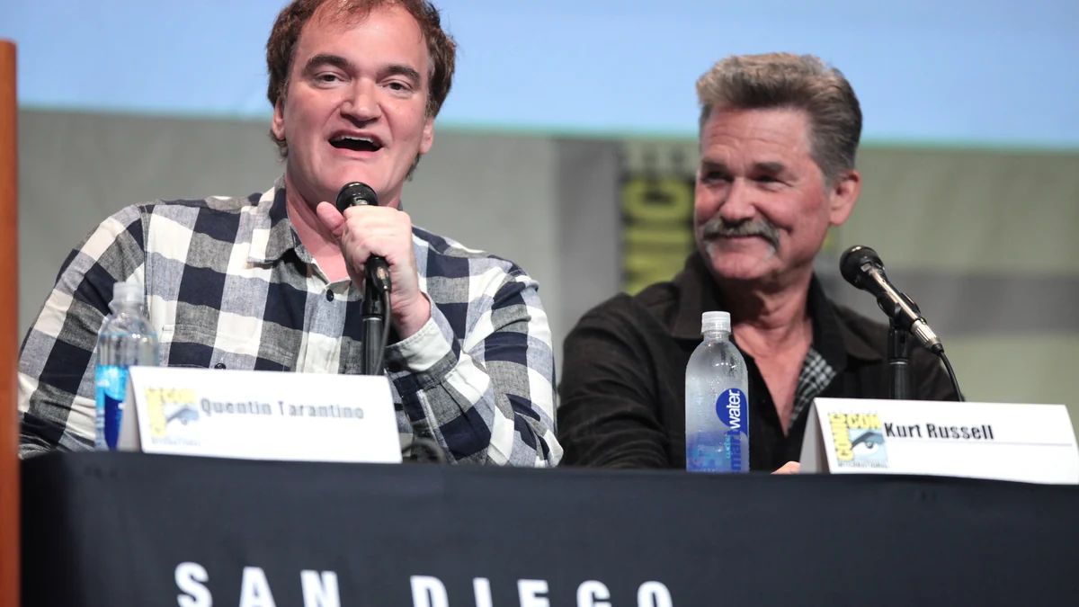 Quentin Tarantino smiles while holding a microphone, seated beside Kurt Russell