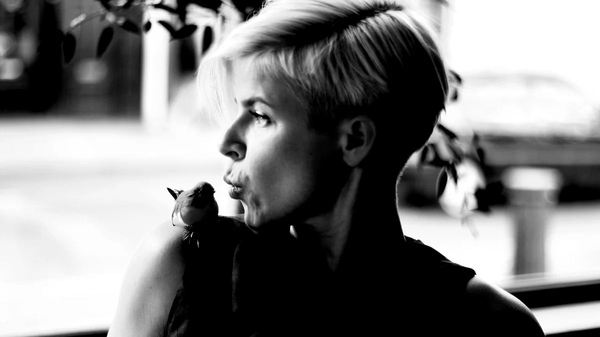 Robyn aparece de perfil en blanco y negro, acercando el rostro a un pequeño pájaro sobre su hombro