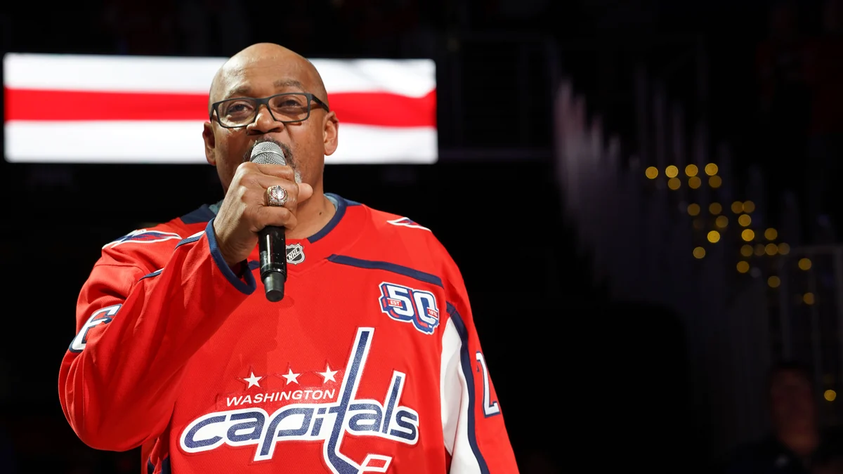 Homem com camisa vermelha dos Washington Capitals canta ao microfone dentro da arena.