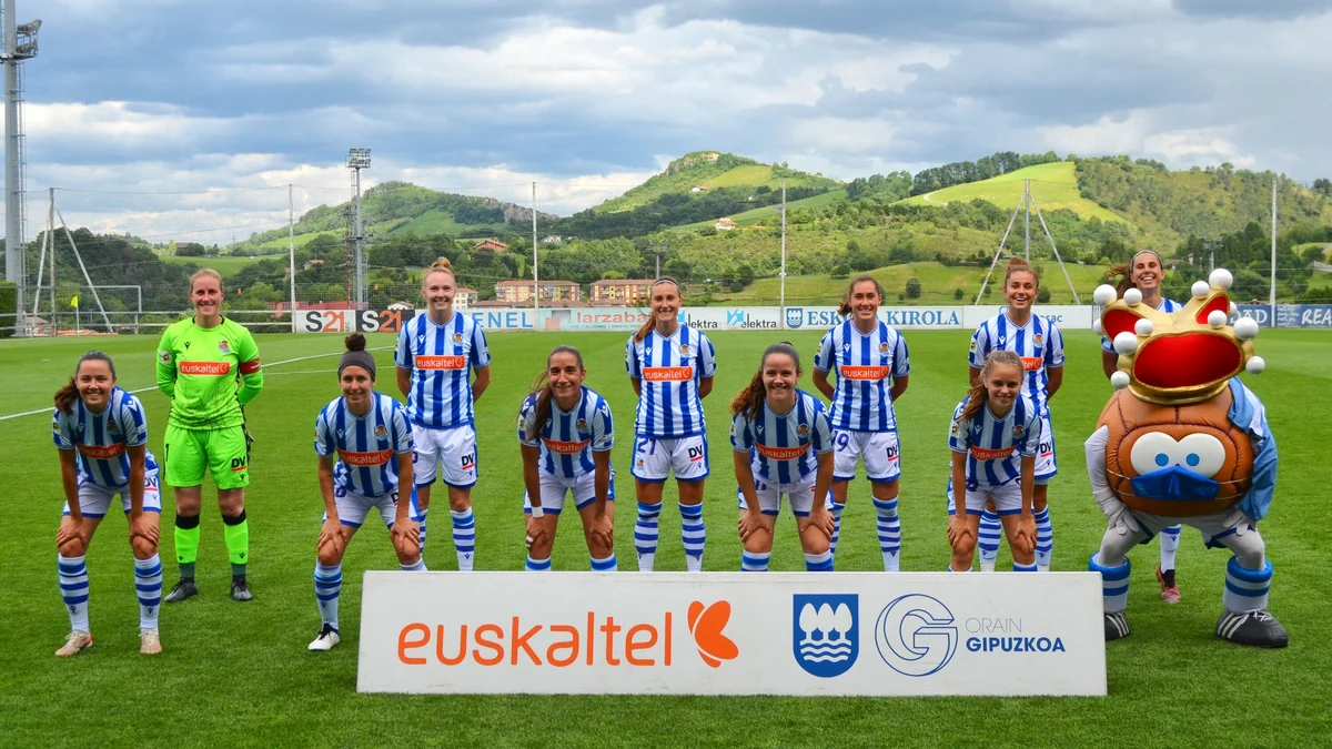 Real Sociedad players pose in two rows on the pitch beside a crowned mascot