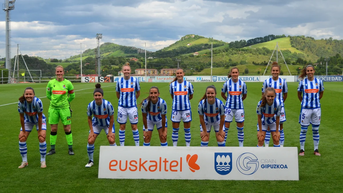 Eleven Real Sociedad players pose on the pitch behind a sign, with green hills in the background