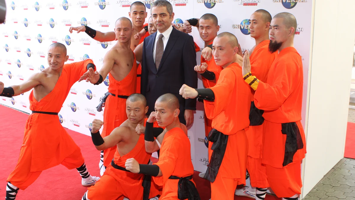 Rowan Atkinson con traje rodeado de artistas vestidos de naranja en una alfombra roja