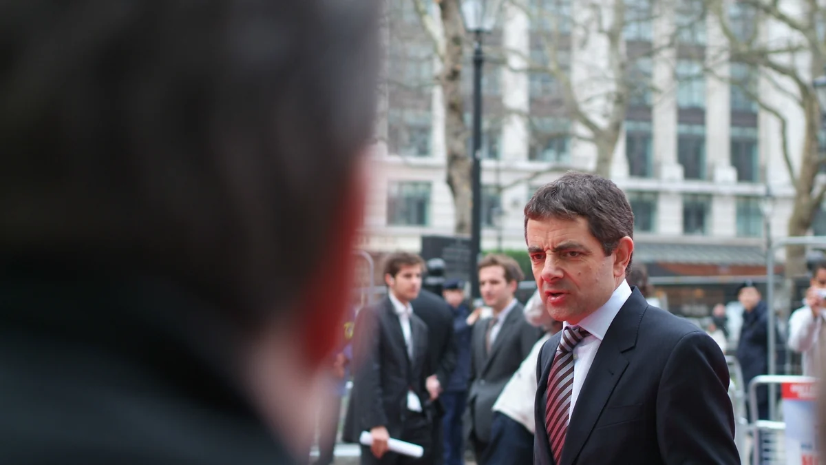 Rowan Atkinson con traje y corbata en un evento al aire libre, con una persona desenfocada en primer plano