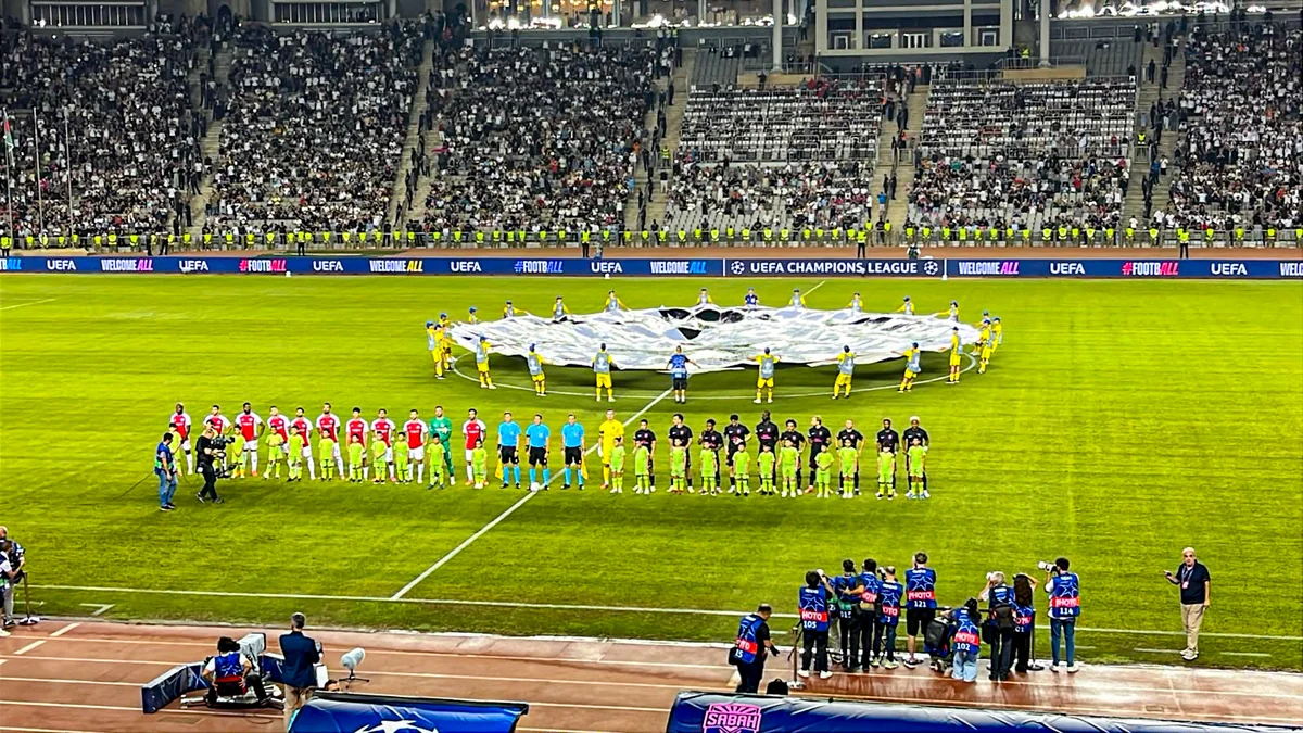 Equipes alinhadas no campo enquanto uma grande bandeira da Champions League é aberta no círculo central diante das arquibancadas cheias.