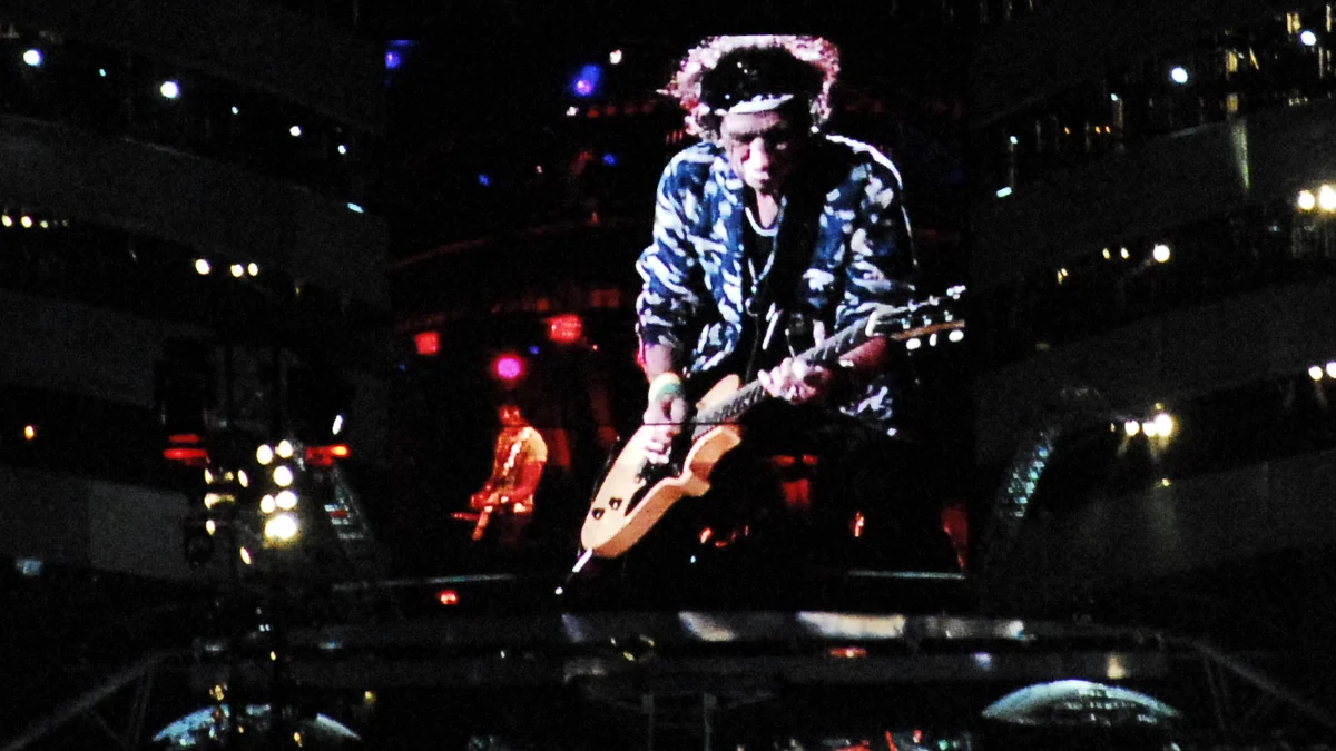 Keith Richards plays guitar on a dark stage, shown on a large screen