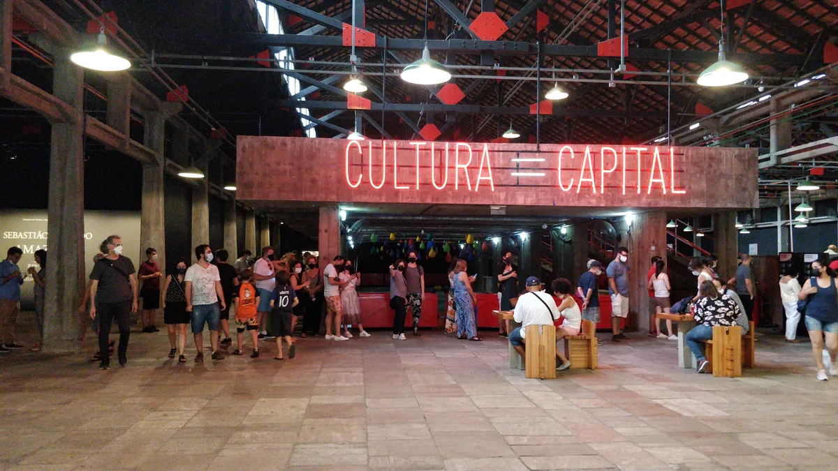 Interior amplo do Sesc Pompeia com estrutura metálica aparente, luminárias suspensas, visitantes e letreiro luminoso Cultura é Capital