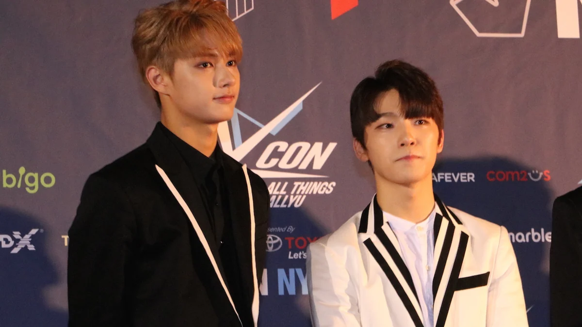 Jun et Dino posent devant un panneau KCON, l’un en costume noir et l’autre en veste blanche