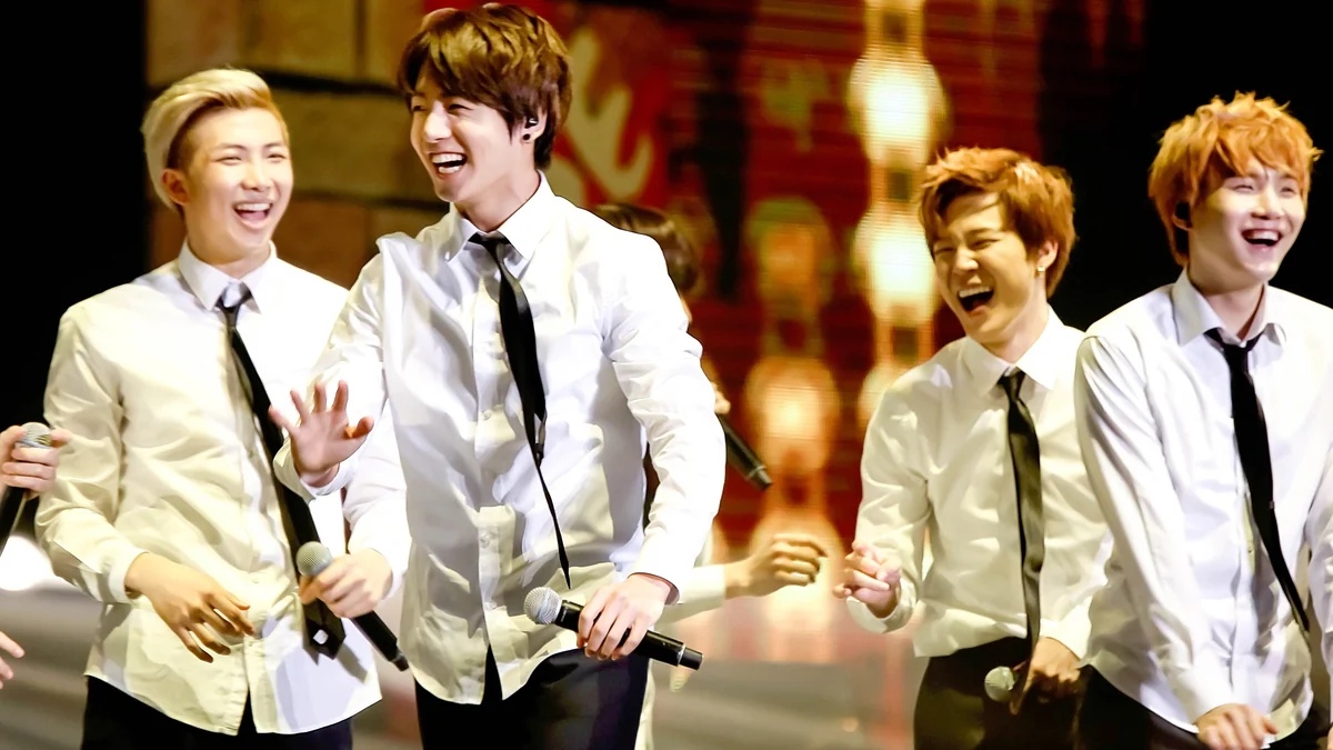 Quatro integrantes do BTS sorriem no palco usando camisas brancas e gravatas pretas