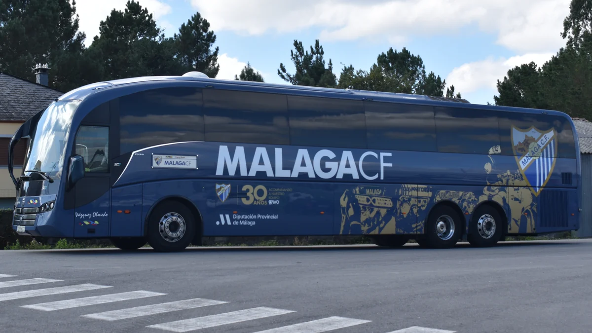 Autocar bleu du Málaga CF garé au bord d’une route, avec le nom et l’écusson du club sur le côté.