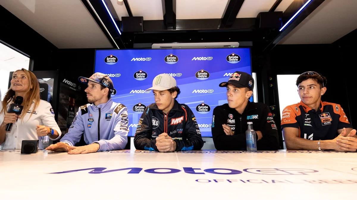 Four riders seated at a table look toward a presenter holding a microphone at a MotoGP event.