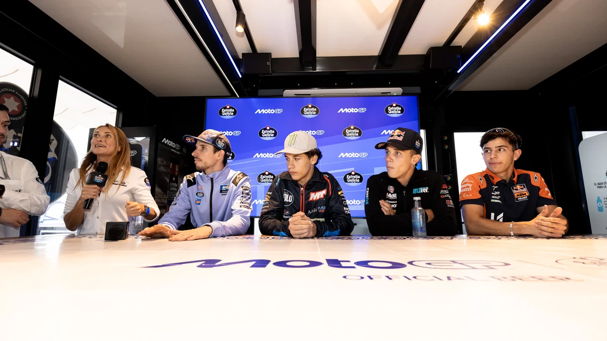 A presenter speaks beside four seated riders in front of a MotoGP backdrop.