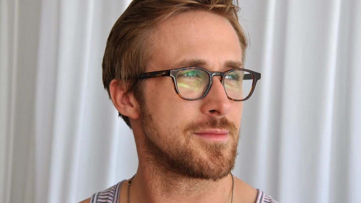 Ryan Gosling de óculos, visto de perfil diante de uma cortina clara