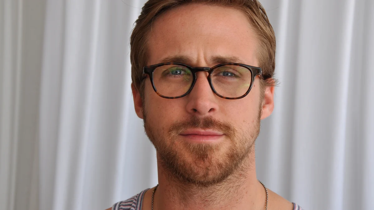 Retrato frontal de Ryan Gosling usando óculos diante de uma cortina clara