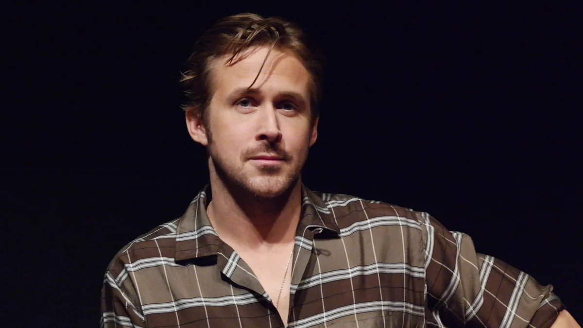 Ryan Gosling sentado no palco, com camisa xadrez e fundo escuro