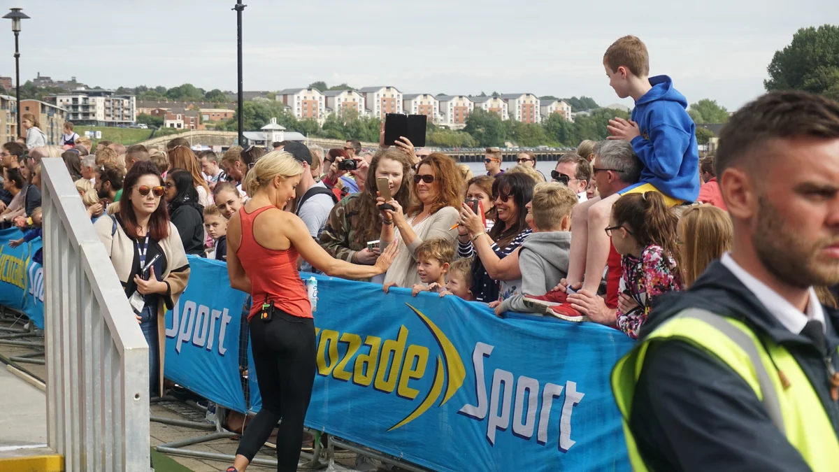 Woman in a red sports top greets spectators behind a barrier at an outdoor event