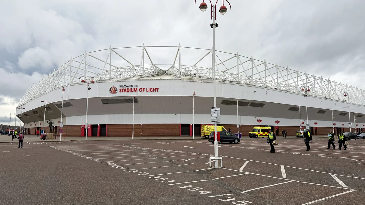 Fachada blanca del Stadium of Light bajo un cielo nublado, con farolas, vehículos y personas en el aparcamiento