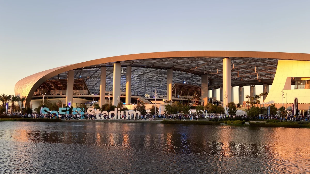Fachada curva del SoFi Stadium al atardecer, con un lago en primer plano