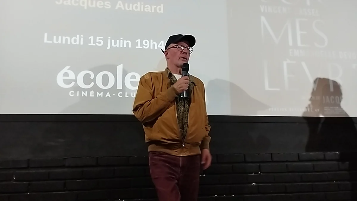 Jacques Audiard sostiene un micrófono ante una pantalla de cine con textos proyectados al fondo.