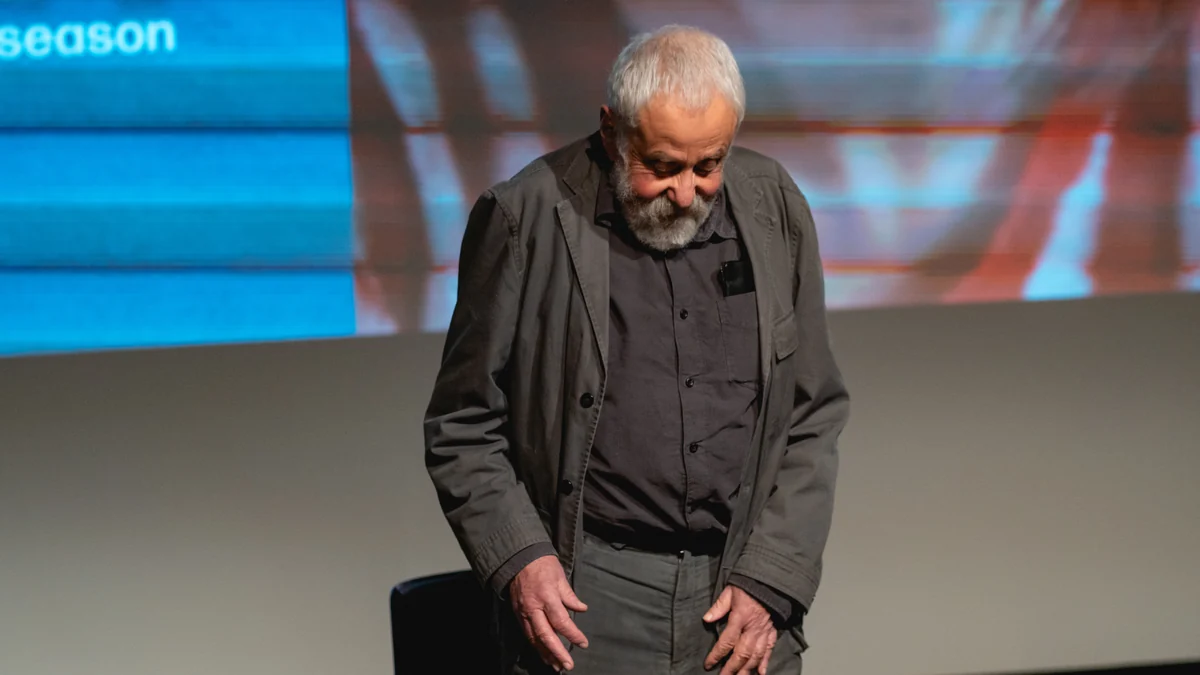 Mike Leigh, with a white beard and dark clothing, lowers his head while standing in front of a screen