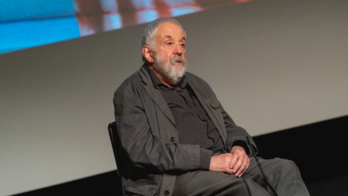 Mike Leigh, with a white beard and dark clothing, seated in front of a screen and looking upward