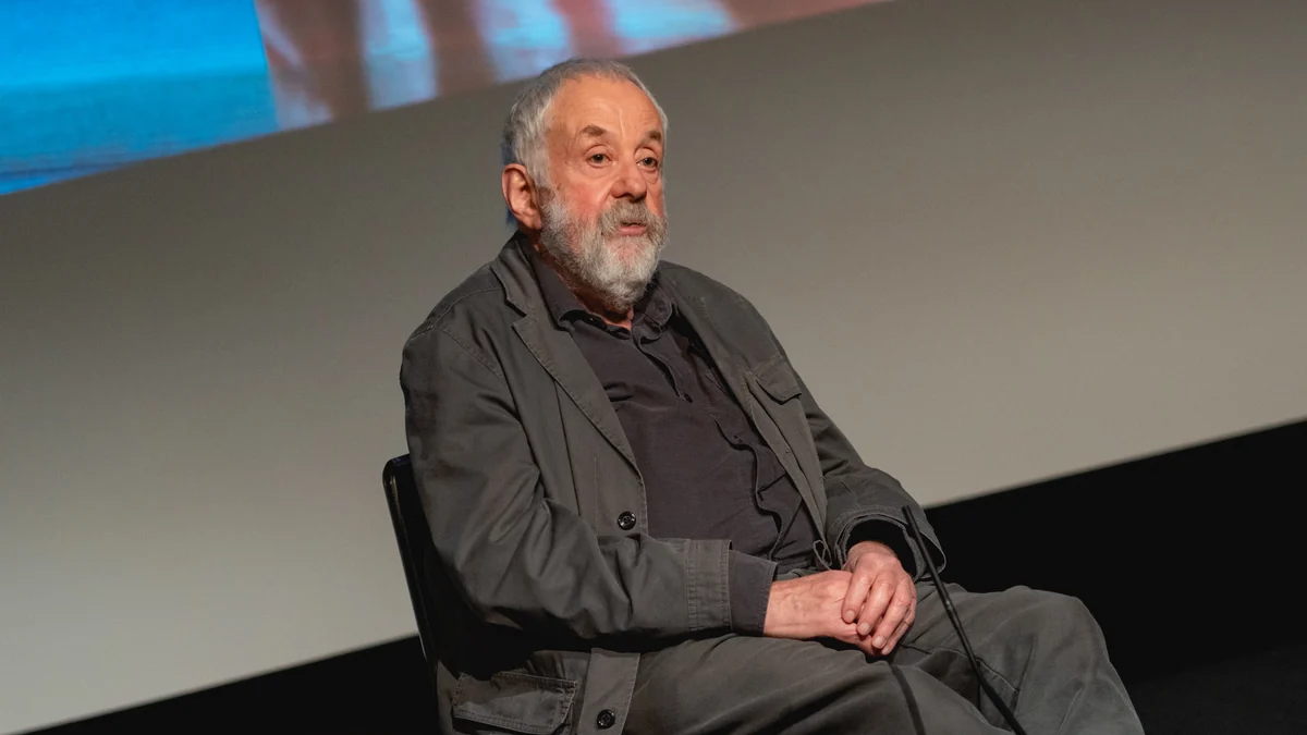 Mike Leigh, with a white beard and dark clothing, seated with his hands clasped in front of a screen