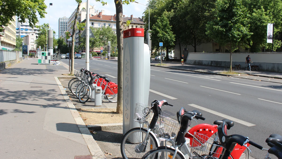 Bicicletas vermelhas e cinzas em uma estação Vélo’v junto a uma avenida arborizada