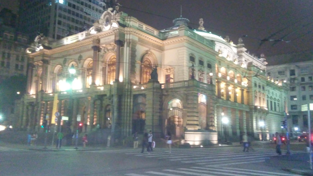 Theatro Municipal iluminado de noche, visto desde una esquina con paso peatonal y personas en la acera.