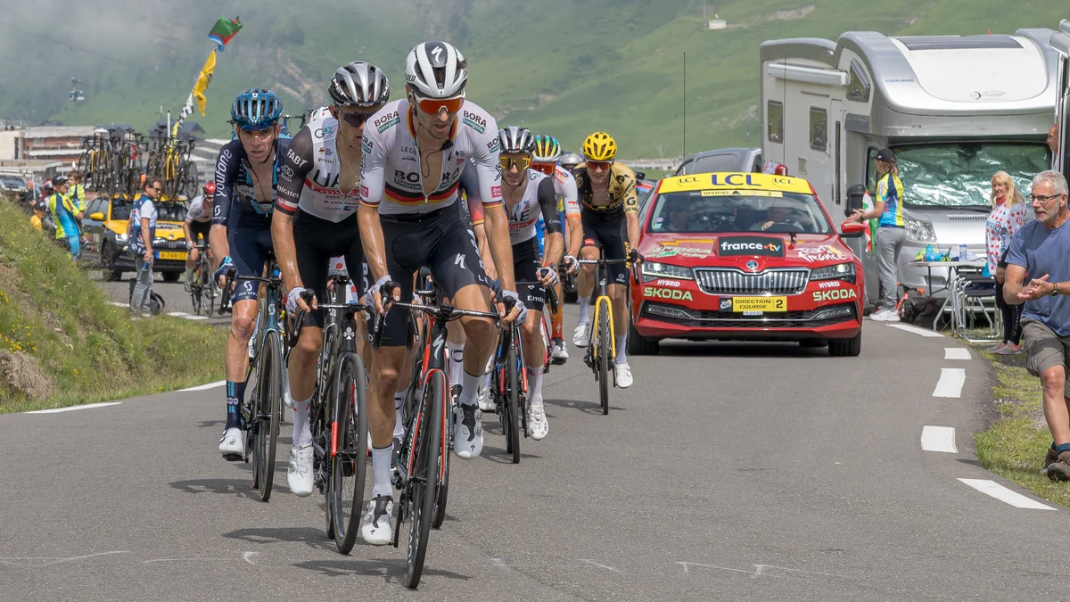 Peloton climbs a mountain road with an official car and spectators alongside
