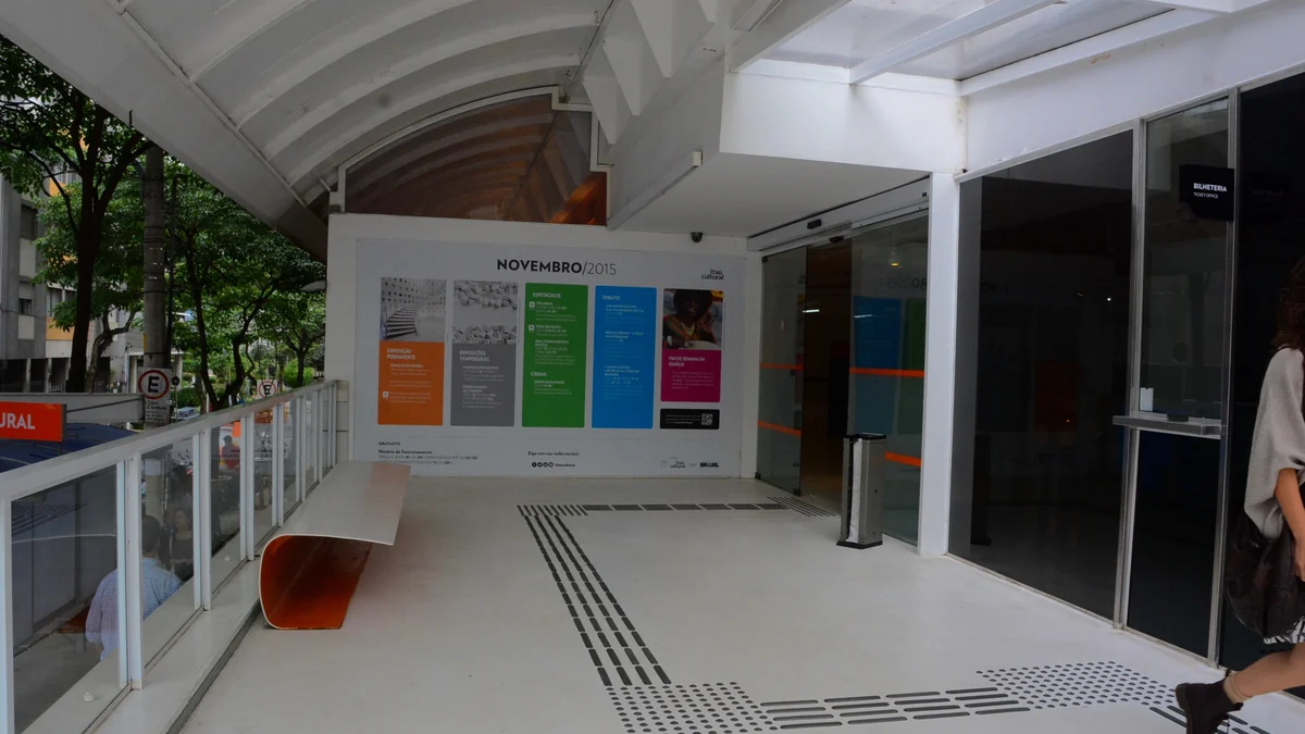 Zona de entrada de Itaú Cultural con rampa, banco, puertas de vidrio y panel de programación