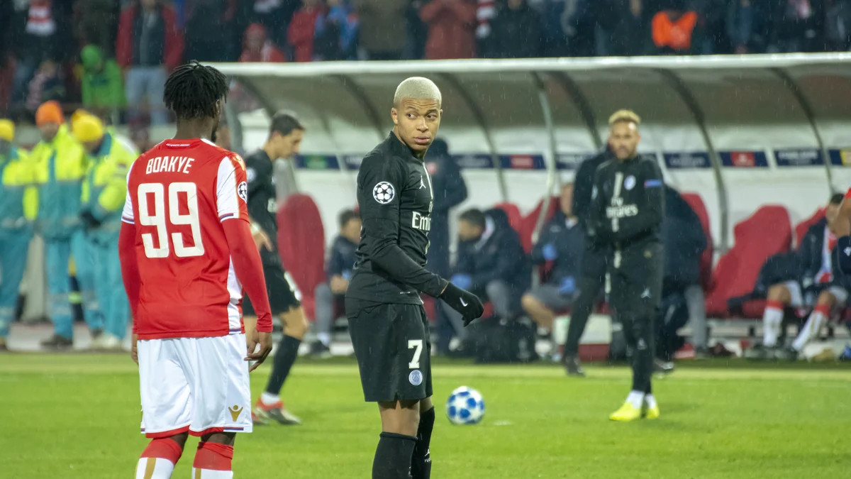 Kylian Mbappé, wearing a black number 7 kit, stands on the pitch in the rain during a football match.
