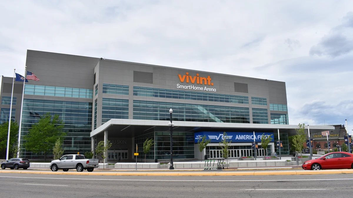 Entrance to Vivint SmartHome Arena, with a glass facade, flags, trees, vehicles and scooters in front.