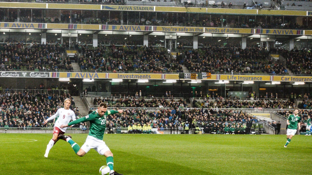 Jogador da Irlanda chuta a bola diante de um adversário da Dinamarca em estádio lotado