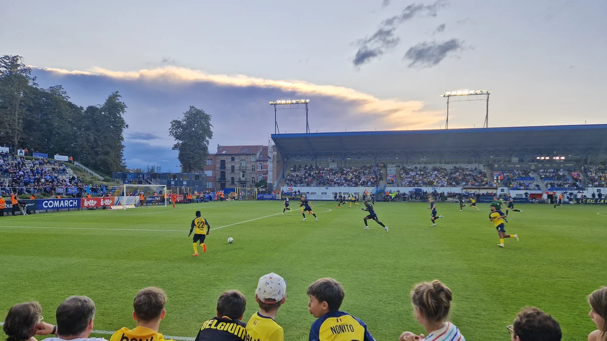 Vista ampla de uma partida de futebol, com jogadores de amarelo e azul em campo e torcedores nas arquibancadas.