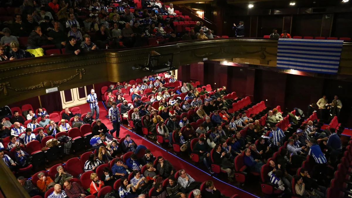 Torcedores da Real Sociedad ocupam poltronas vermelhas em um teatro, com uma bandeira azul e branca no balcão.