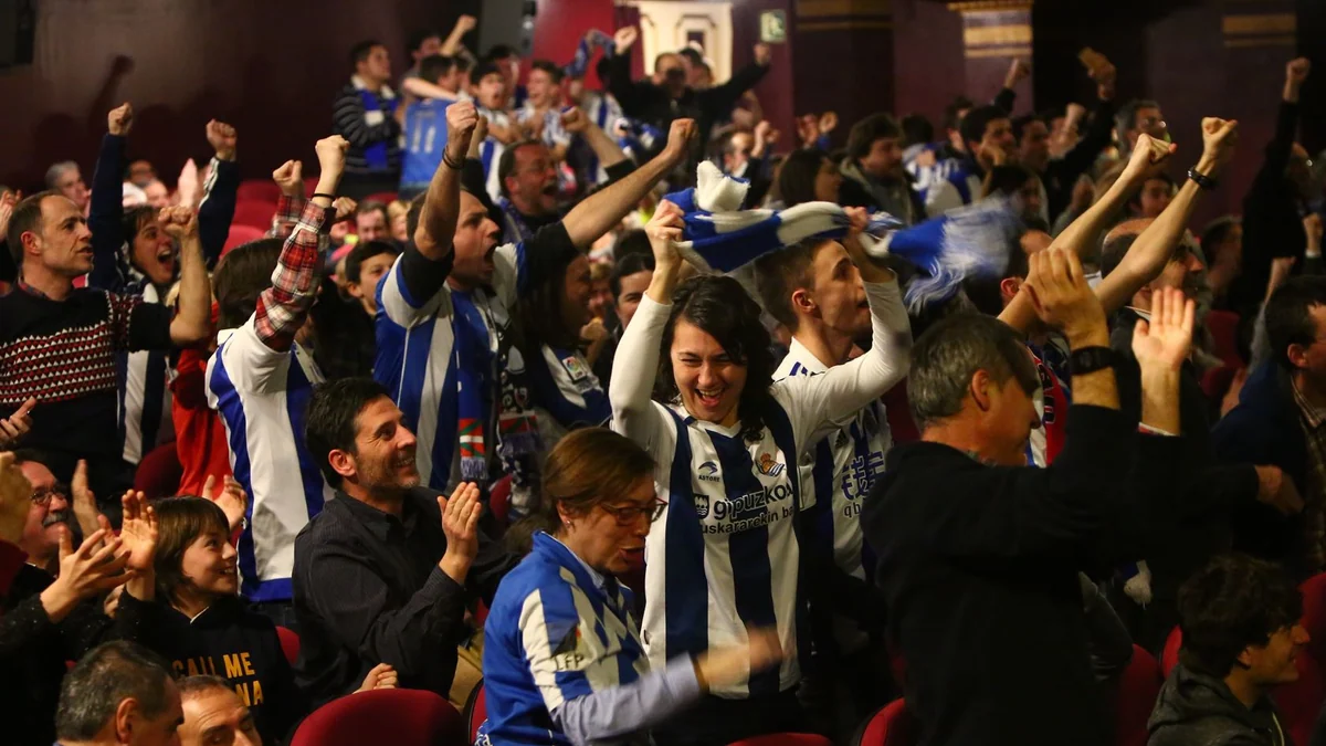 Torcedores da Real Sociedad comemoram de braços erguidos entre poltronas vermelhas de um teatro.