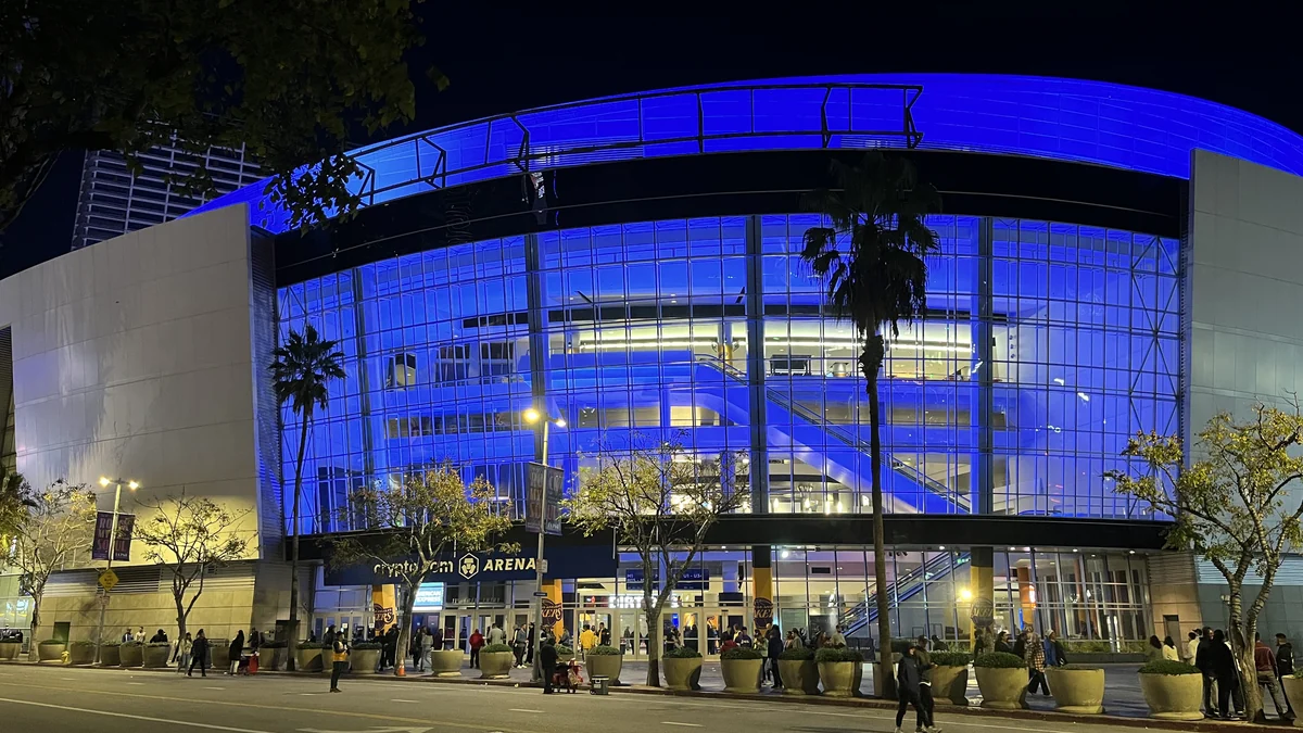 Curved glass facade of Crypto.com Arena illuminated in blue at night, with palm trees and people on the sidewalk