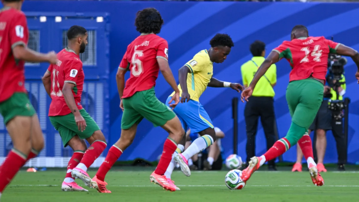 Vini Jr., com o uniforme amarelo do Brasil, avança entre três jogadores de Marrocos vestidos de vermelho e verde.