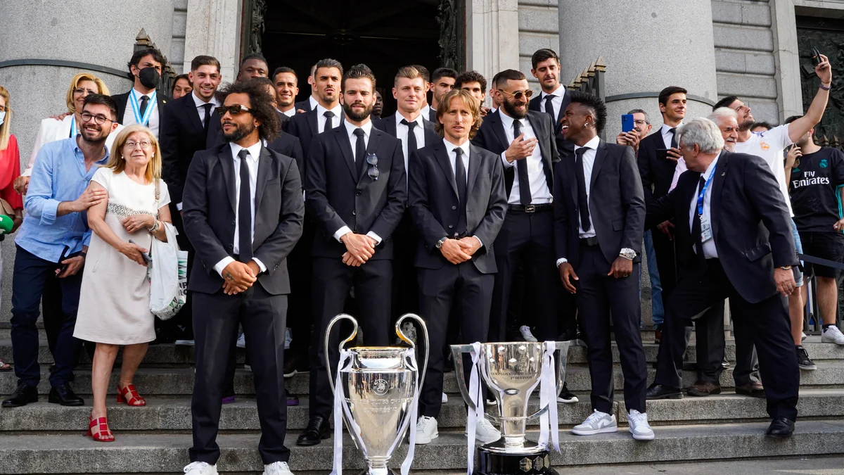 Des joueurs du Real Madrid en costume posent sur un escalier avec deux trophées ; Vinícius Júnior se tient à droite.