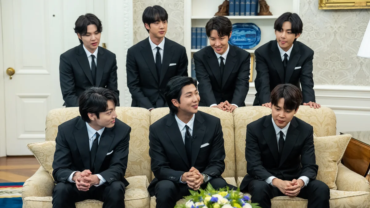 Sete integrantes do BTS, de ternos pretos, reunidos diante de um sofá em uma sala clara.