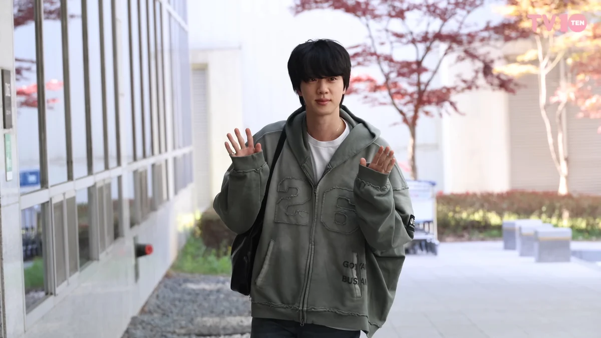 Jin, de cabelo preto e moletom cinza, acena com as duas mãos em uma área externa.
