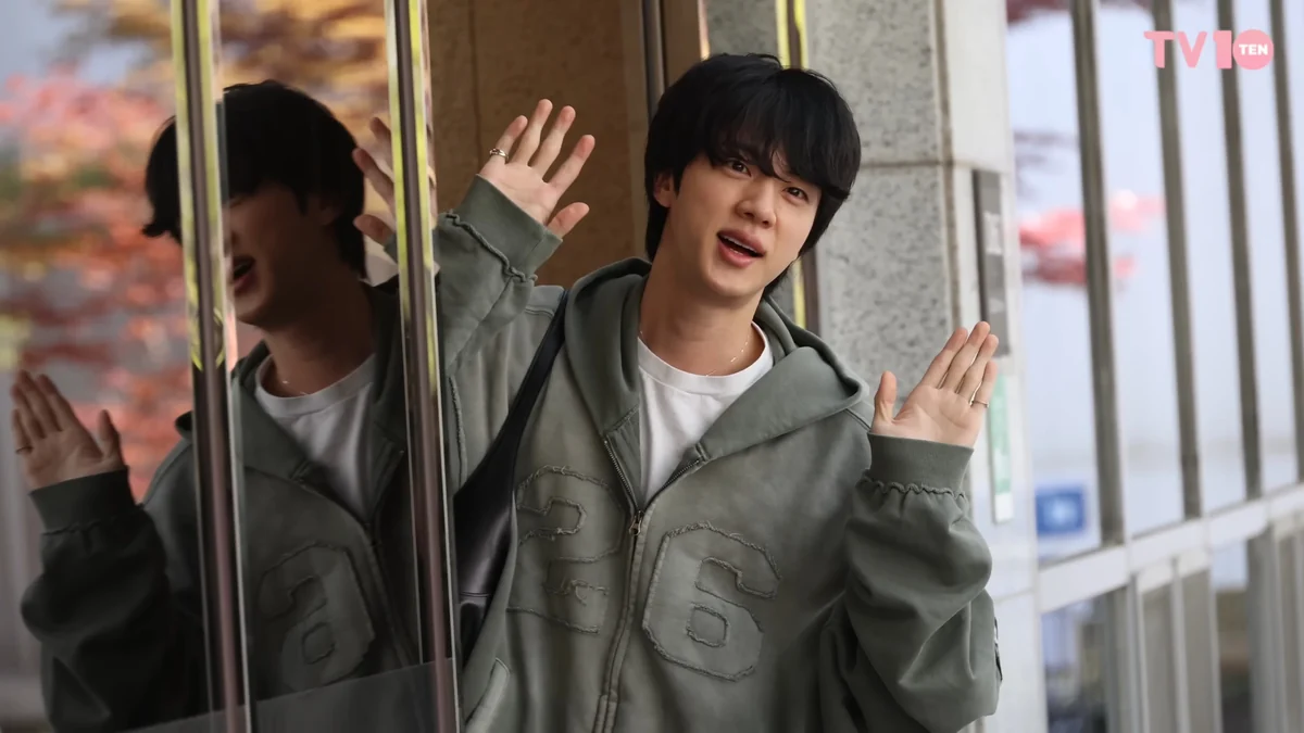 Jin, com moletom cinza, acena com as duas mãos ao lado de uma porta de vidro que reflete sua imagem.