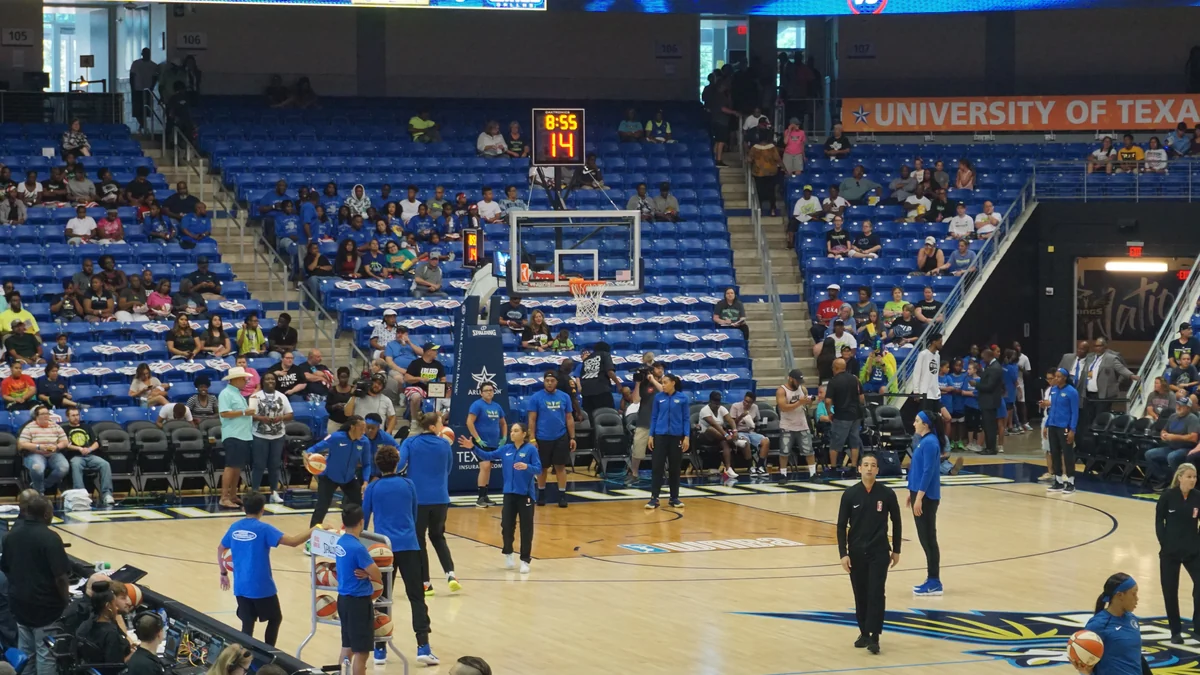 Jogadoras e integrantes da equipe de azul aquecem perto da cesta em uma quadra de basquete.