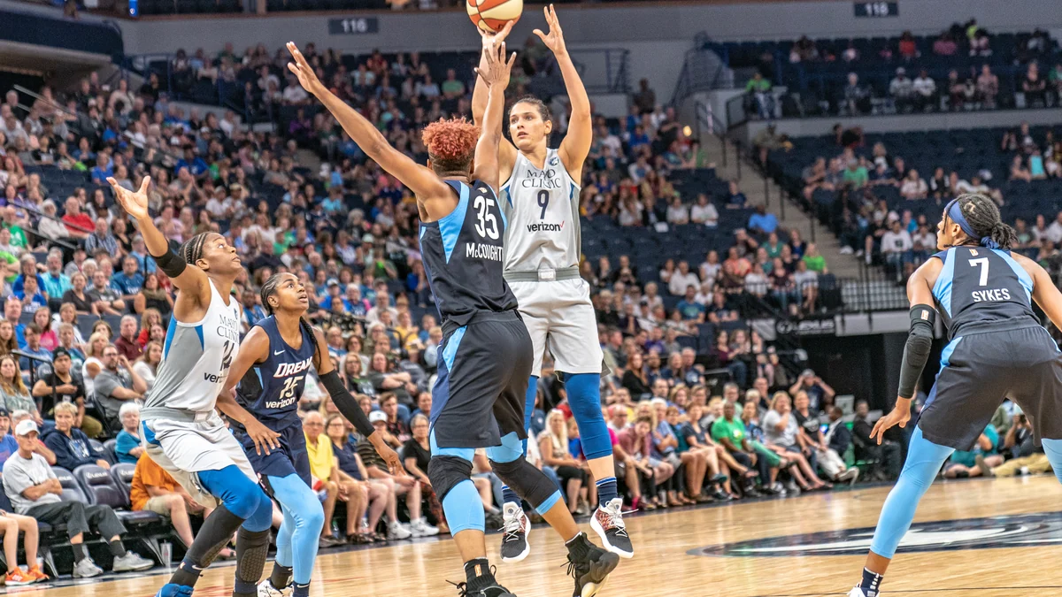 Una jugadora de Minnesota Lynx lanza sobre una defensora de Atlanta Dream, rodeada de otras jugadoras y del público.