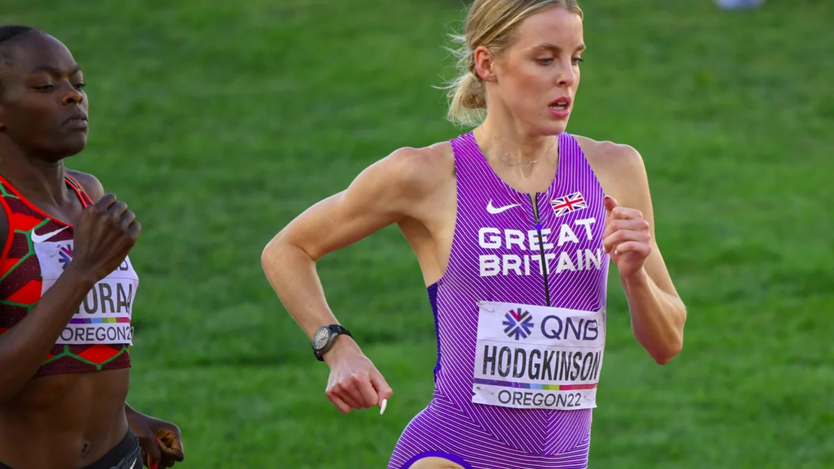 Atleta britânica com uniforme roxo e número Hodgkinson corre ao lado de outra competidora.