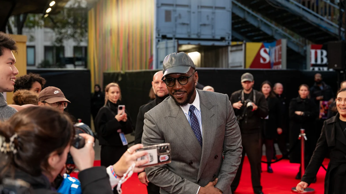 Mahershala Ali con traje gris inclinado hacia seguidores junto a una alfombra roja
